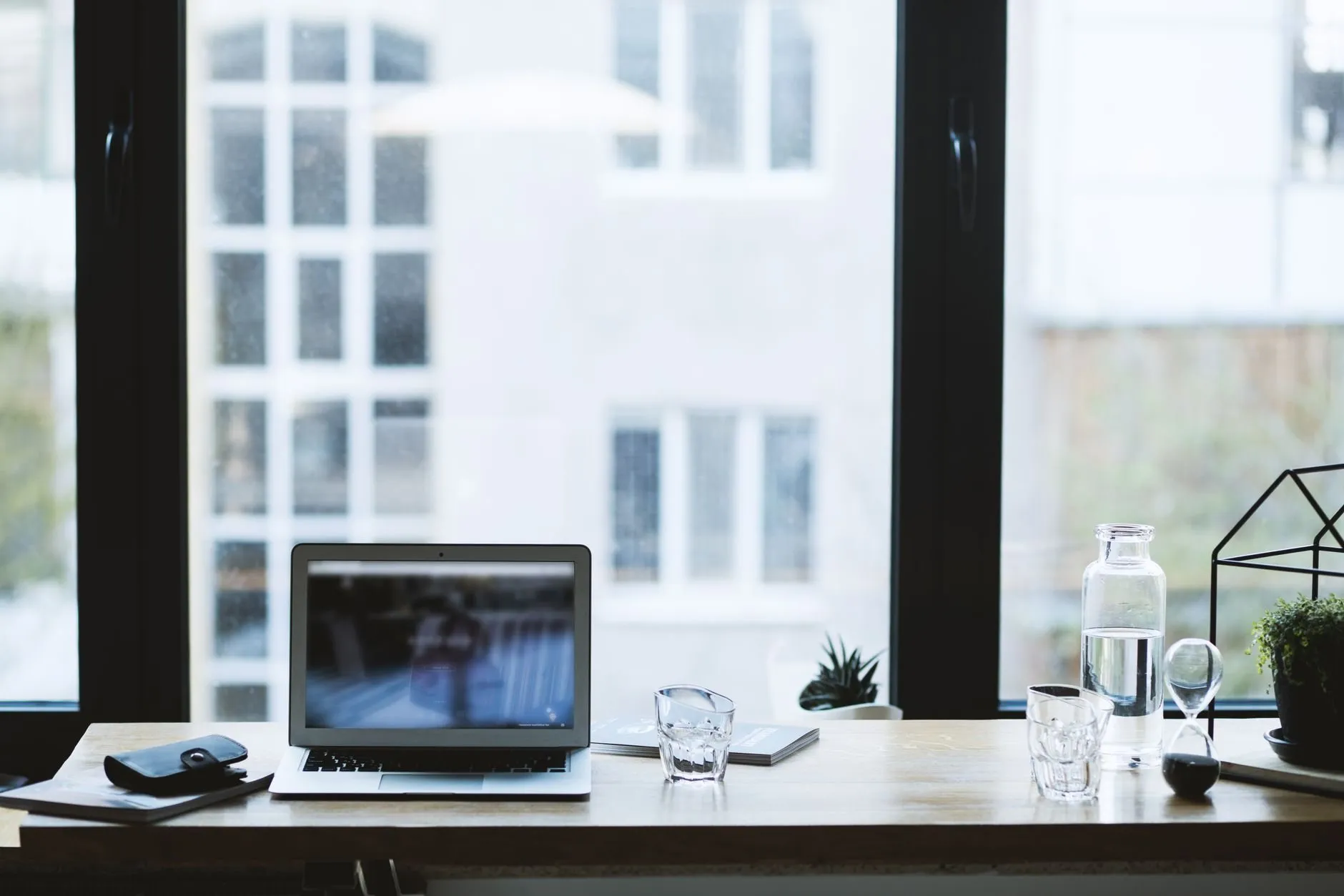 A quiet desk by a tall window with a laptop, a water glass and a small plant