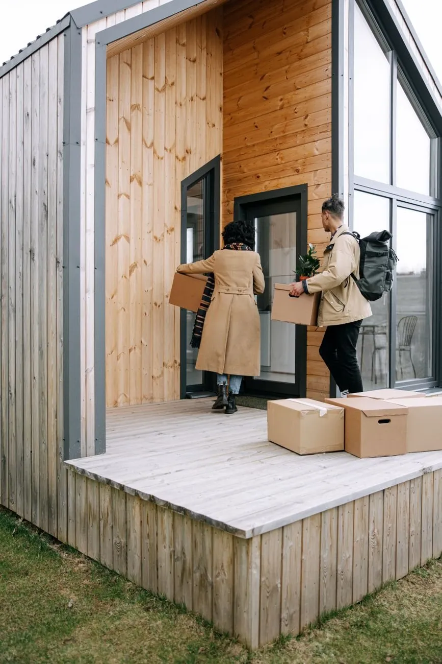 Two people carrying cardboard boxes and a plant into a timber-clad house