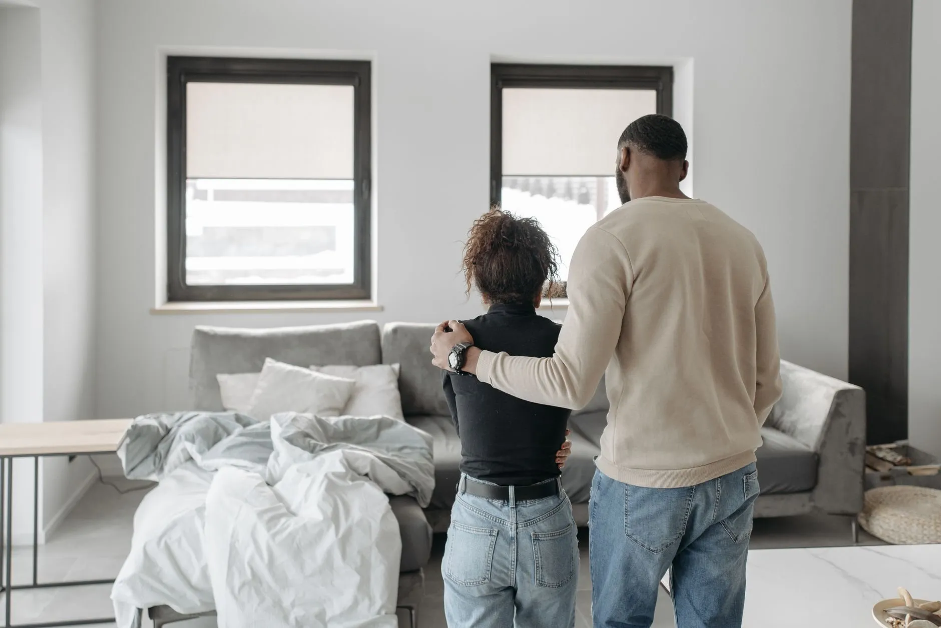 A couple seen from behind in a bright living room, an arm around a shoulder