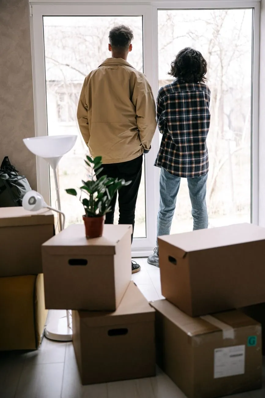 A couple seen from behind at a tall window, moving boxes stacked around them