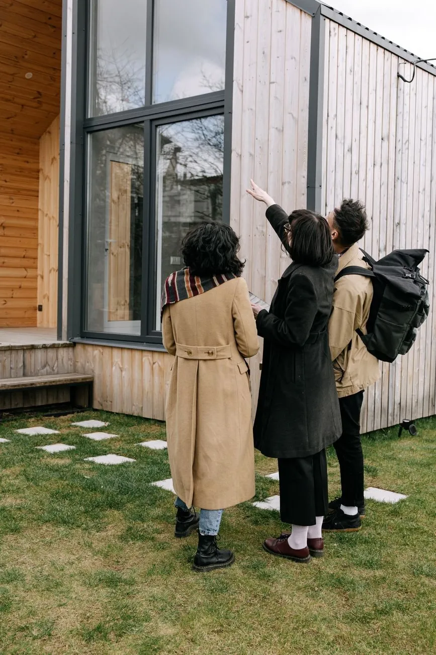 Three people seen from behind outside a timber-clad house, one pointing up at the windows